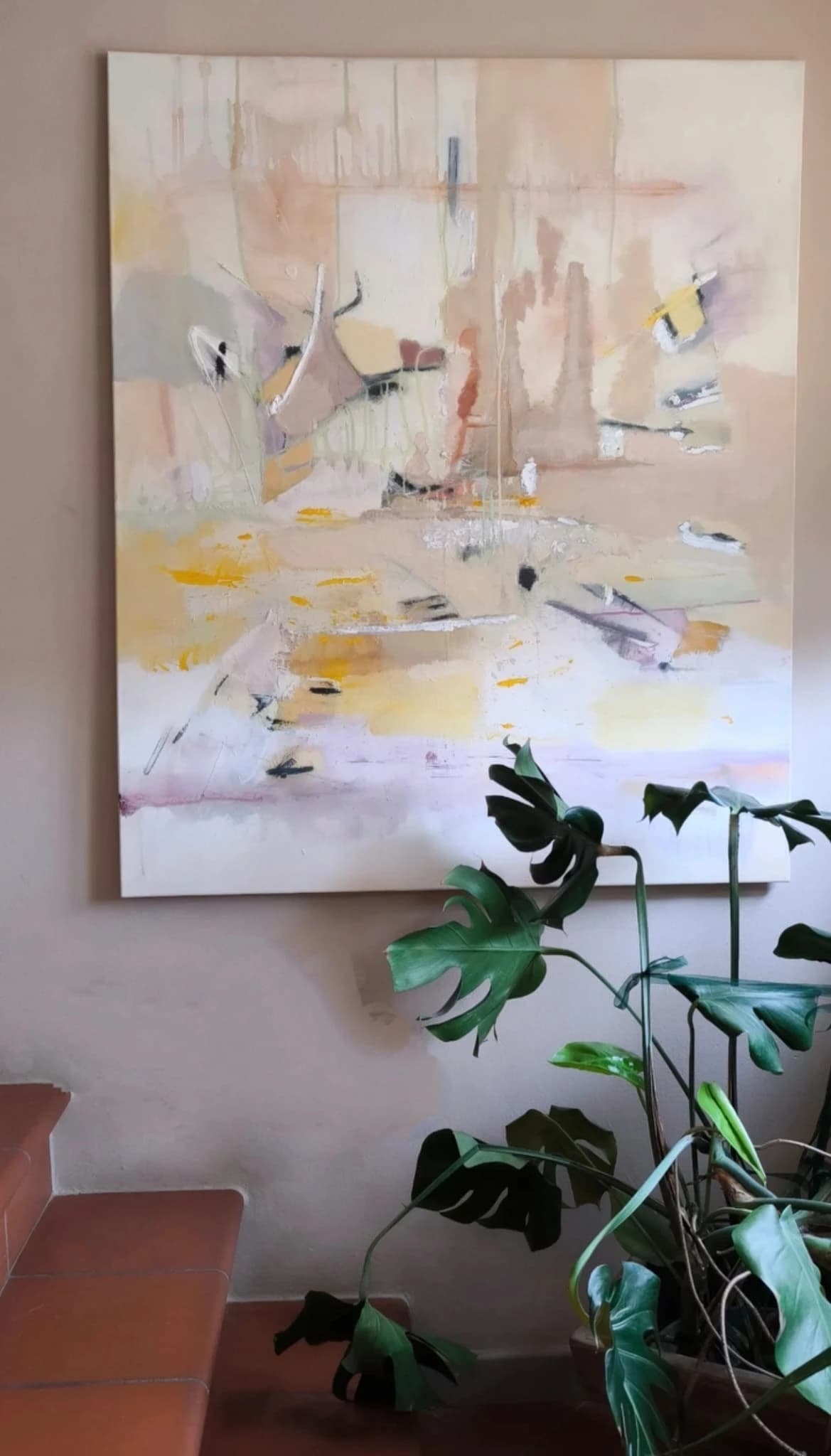 A large pale ochre painting on a warm plaster wall, beside a monstera and terracotta steps